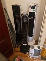 Three tower fans including two tall black fans and a white fan with control panel on top standing in a corner. White Honeywell humidifier and metal folding step ladder next to fans. Items untested and stored indoors.