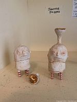 Front view of two ceramic sculptures and detached headpiece of one sculpture showing painted faces and striped legs.
