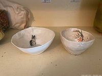 Two white, free-form art pottery bowls with painted rabbit and floral designs inside, placed side by side on a shelf.