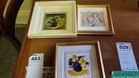 Photo showing three small framed artworks on a wooden table: sunflower painting in thick off-white frame, a small colorful abstract sketch, and a small floral watercolor painting.