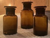 Three amber apothecary jars shown from front with lids on, with light reflecting through the glass showing amber color.