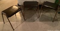 Three mid-century stackable stools with brown vinyl seats and metal legs seen from front and side.