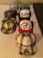 Seven hats arranged in two rows on a surface, different styles and colors with visible logos and patterns.