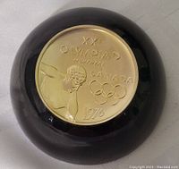Top-down photo of the 1976 Montreal Olympics commemorative paperweight showing the embossed gold-toned medallion with discus thrower, Olympic rings, and text.