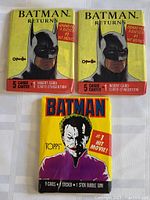 Three unopened Batman movie themed trading card packages on a white surface: two Batman Returns packs and one Topps Joker pack.