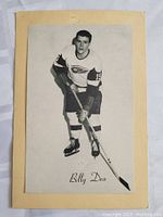 Photo of original vintage black and white hockey photo card of Billy Dea wearing Detroit Red Wings uniform with jersey number 15, mounted on tan paper with printed name 'Billy Dea' at bottom.