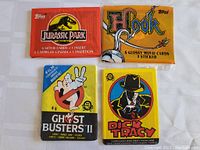 Four unopened vintage trading card packages from movies: Jurassic Park, Hook, Ghostbusters II, Dick Tracy are shown together on a white cloth background.