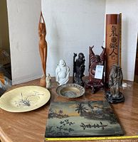 Full view of Asian art collection on table, showing bamboo platter, different wooden carvings, ceramic Buddha, brass plate, and painting.