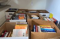 Photo showing 8 large cardboard boxes filled with books in various sizes and colors, arranged on a floor near stairs.