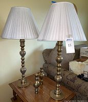 Full view of two brass table lamps on a wooden table with off-white pleated cloth shades, showing the lamps' height and shape.