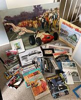 Image showing the large race car painting, Woodies on the Wharf poster, several magazines and books, and framed license plates arranged on the floor.