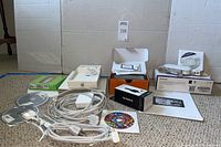 Full lot overview showing Garmin GPS, multiple Apple cables, iPod Shuffles box, Netgear modem box and manual, disc and power cables laid out on floor.