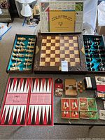 Full overview of box chess set with open wooden carved chess pieces beside decorated carved chessboard, The Game of Denali-opoly board game visible, multiple decks of playing cards, metal Fifteen Puzzle and other game items on table.