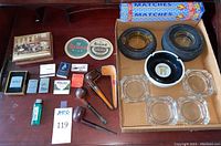 Assortment of ashtrays, pipes, lighters, matchbooks, coasters, and matches arranged on a surface.