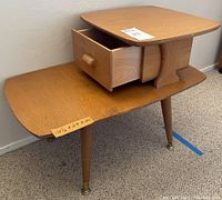 Side view of the mid-century two-level wooden side table with small pull-out drawer on the upper level and tapered legs with brass caps, showing overall condition and surface wear.