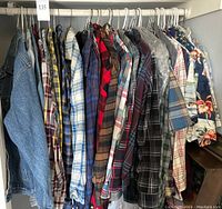 Full view of many men's shirts on hangers, showing a variety of plaid patterns and colors, including flannel fabric shirts and a few Hawaiian shirts.