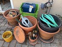 Full lot of terracotta planters, ceramic pot, hose, watering can, crates, and gardening tools arranged on paved ground.