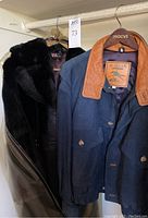 Two coats hanging: a black simulated fur coat with leather details, mid-calf length, and a blue lamb nubuck leather and satin jacket with brown leather collar, M. Julian brand.