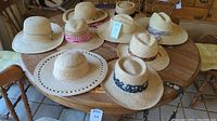 12 men's straw hats displayed on a wooden round table in a room with wooden furniture. Hats feature various cloth hatbands and natural woven straw.