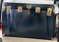 Front view of a large black travel trunk with brass latches, locks, corner reinforcements, and studded edge trim.