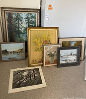 Seven pieces of wall art arranged, including forest scene, floral paintings, windmill winter scene, and black and white print on beige carpeted floor