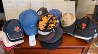 Over 40 worn baseball caps and 5 other hats displayed on wooden furniture, showing variety of logos and styles including San Francisco Giants and Viking River Cruises.