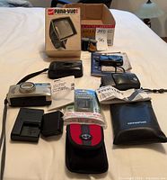 Wide shot showing the assortment of cameras and accessories on a white surface including black Olympus Infinity Mini camera with case, silver Nikon camera, Sony Cyber-shot, Gaf Pana-Vue 1 viewer, charger, film rolls, camera cases.