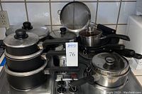 Full view of various pots, pans including cast iron skillets and Circulon skillets stacked on stovetop with a large sieve behind.
