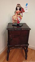Full view of wooden humidor table with closed door, Japanese doll standing on top.