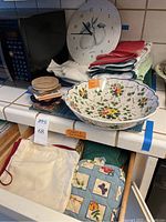 Photo showing the Italian pasta bowl, assorted kitchen towels and potholders, mixed coasters, and cloth items arranged on a kitchen counter near a microwave.