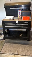 Full view of the black metal Kobalt workbench with three drawers and upper cabinet, with various tools and boxes on top and a cushioned rug underneath