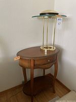 Gold tone lamp with circular glass shade on oval wooden table with one drawer and lower shelf