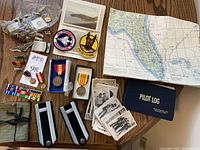 Overview photo showing assorted military pins, medals, patches, photographs, folded map, and pilot log books on a wooden table.