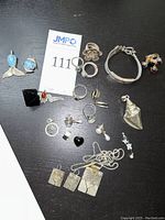 Overall view of assorted silver jewelry items laid flat on black surface including pendants, rings, bracelet, earrings and pin.