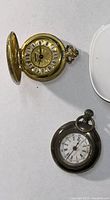 Top open view of gold-tone pocket watch showing Roman numeral dial and ornate crown, alongside closed silver-tone pocket watch with worn dial.