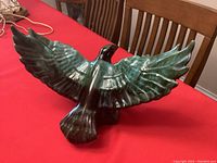 Front and top side view of blue-green glazed ceramic eagle/hawk figurine with widely spread wings on a wooden table with red tablecloth.