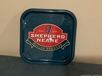 Square metal sign with dark blue background and a red shield emblem with lettering for Shepherd Neame Master Brewers.