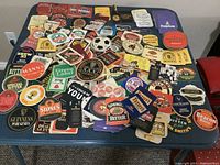 Photo showing an arrangement of approximately 80 assorted British beer and drink mats and coasters of varying shapes and colors laid on a table.