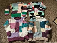 Four hand knit sweaters laid out on carpet floor, showing front and back patchwork color block design.