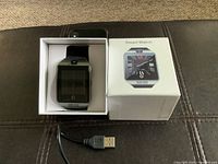 Smart watch displayed inside original box with lid beside it and USB charging cable below.