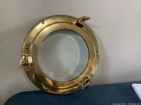 Front side of an 11 1/4 inch round solid brass porthole with glass door, polished brass with three clasps, clear glass in center.