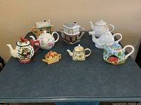 Full view of 9 decorative teapots including holiday-themed, fruit basket-shaped, and house-shaped teapots.