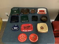 View of all thirteen British beer ashtrays arranged on table in various materials including glass, plastic melamine, and metal, different shapes and colors with distinct logos.
