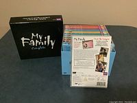 Photo showing the black 'My Family Complete' slipcase box along with the blue box containing the set of DVDs vertically arranged. One DVD case is taken out and shown with back label visible.