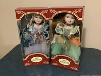 Photo of two boxed porcelain dolls, one with red hair and the other blond, each 16 inches tall, dressed in decorative dresses with floral and ribbon details.