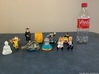 Full view of the ten toy items including the sailor captain, ship in bottle, ET figure, Disney characters (Sylvester and Tweety), McDonald's toys, and others. A Coca-Cola bottle placed for size reference.