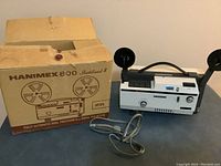 Photo of the Hanimex 800 projector with original box showing the unit with reel arms and power cord on a flat surface.