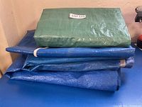 Stack of four tarps, three blue and one green, folded neatly with visible texture and edges, showing the four tarps included in the lot.