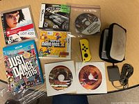 Top-down image showing assorted video games and accessories on a wooden surface including game cases for Wii U, Nintendo DS, game discs in paper sleeves, a yellow Joy-Con controller and VR headset.
