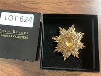 Front view of gold-tone Joan Rivers Classic Collection brooch in starburst design with central oval amber stone and surrounding rhinestones in black box.